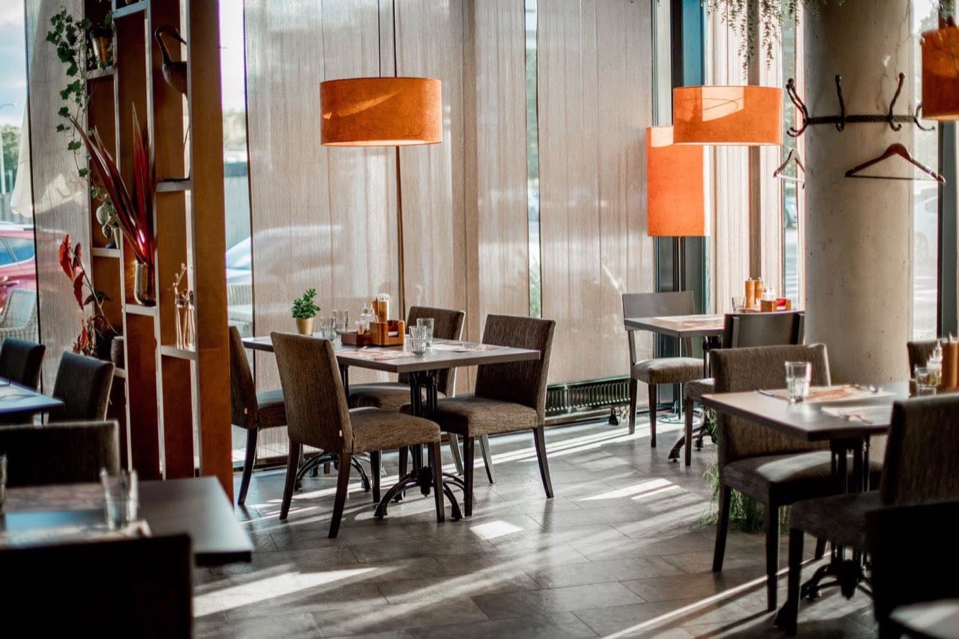 Warmly lit modern restaurant dining room with set tables, upholstered chairs, and orange pendant lamps.